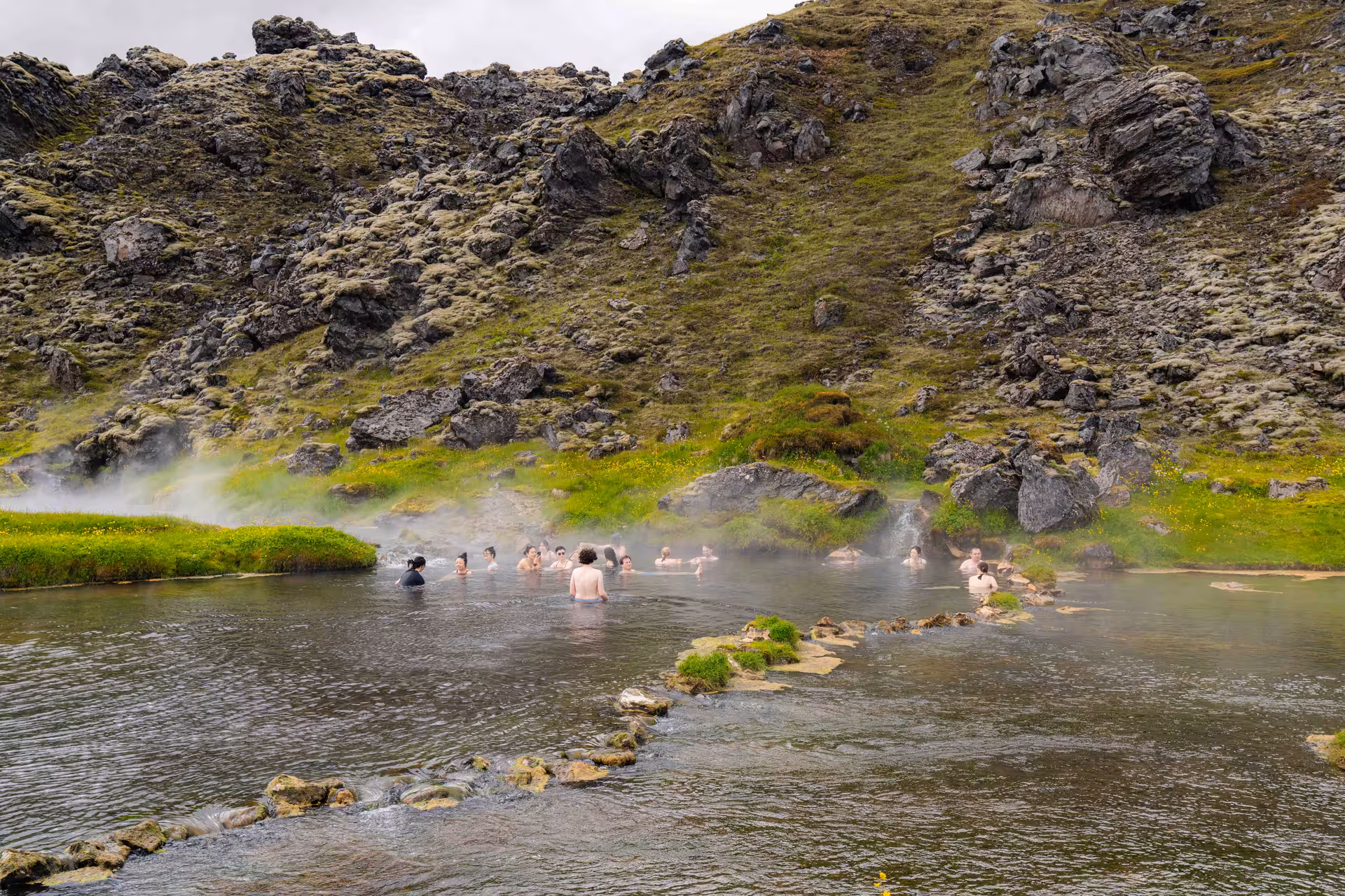 Hikers soaking in steaming Landmannalaugar hot spring river surrounded by mossy lava fields in Iceland