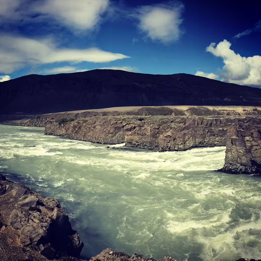 Turbulent glacial river rapids flowing through rocky canyon with dark volcanic mountain in Iceland highlands