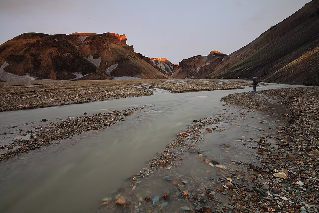 Glacial river winding through Landmannalaugar rhyolite mountains at sunset with lone hiker standing on rocky riverbank