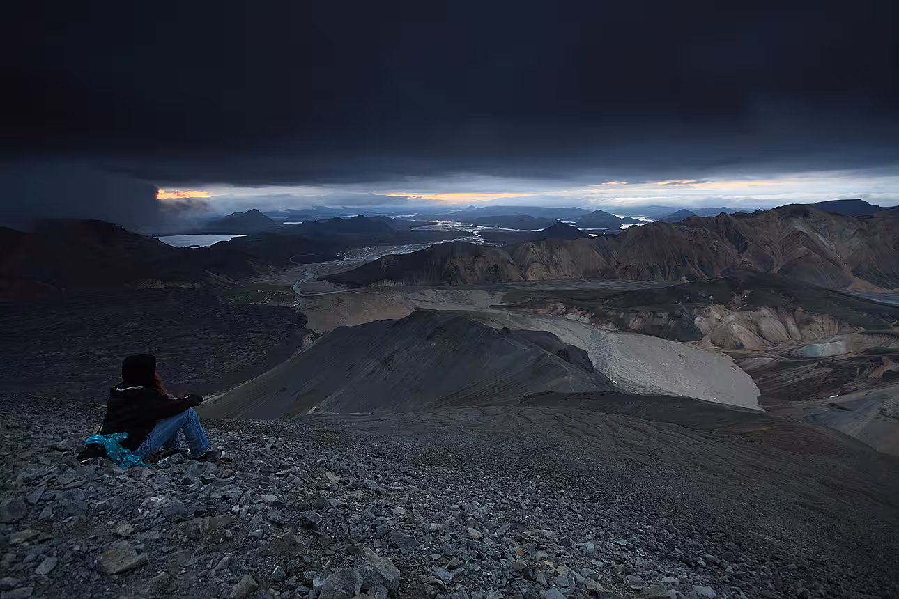 Hiker sitting on rocky ridge overlooking Landmannalaugar rhyolite mountains and river valley under stormy sky