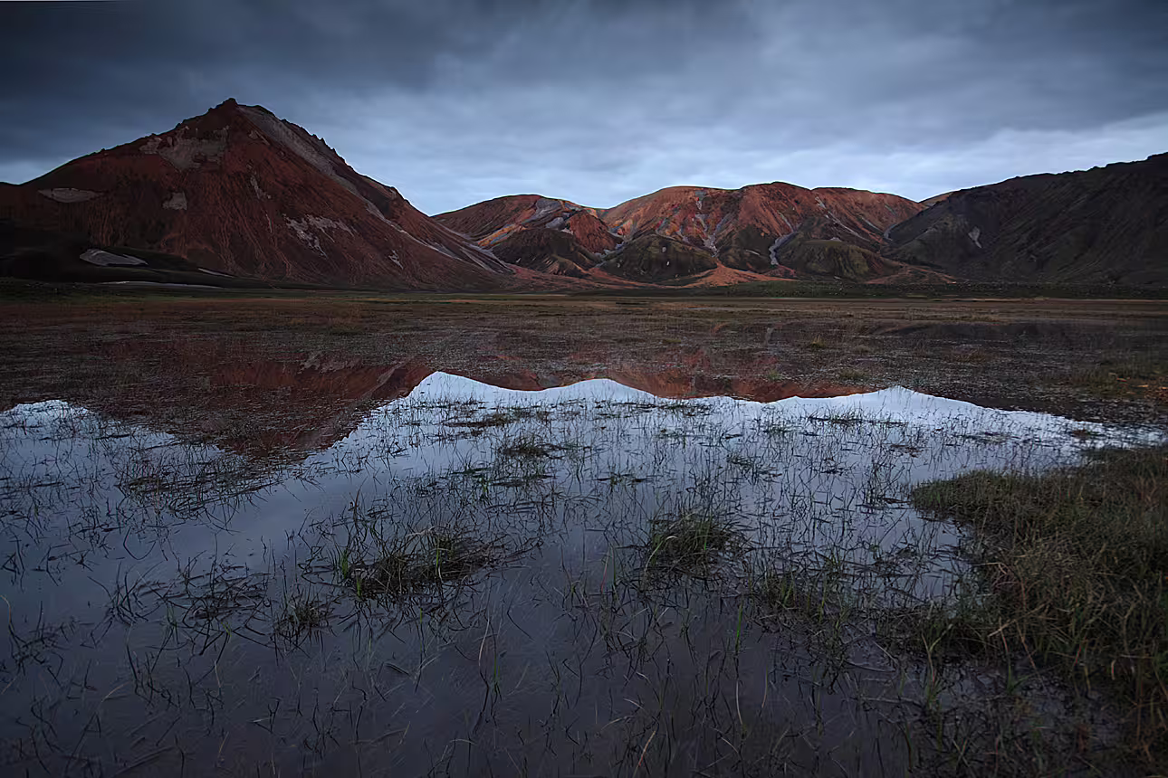 Red rhyolite mountains of Landmannalaugar reflected in a still flooded plain under dramatic cloudy skies