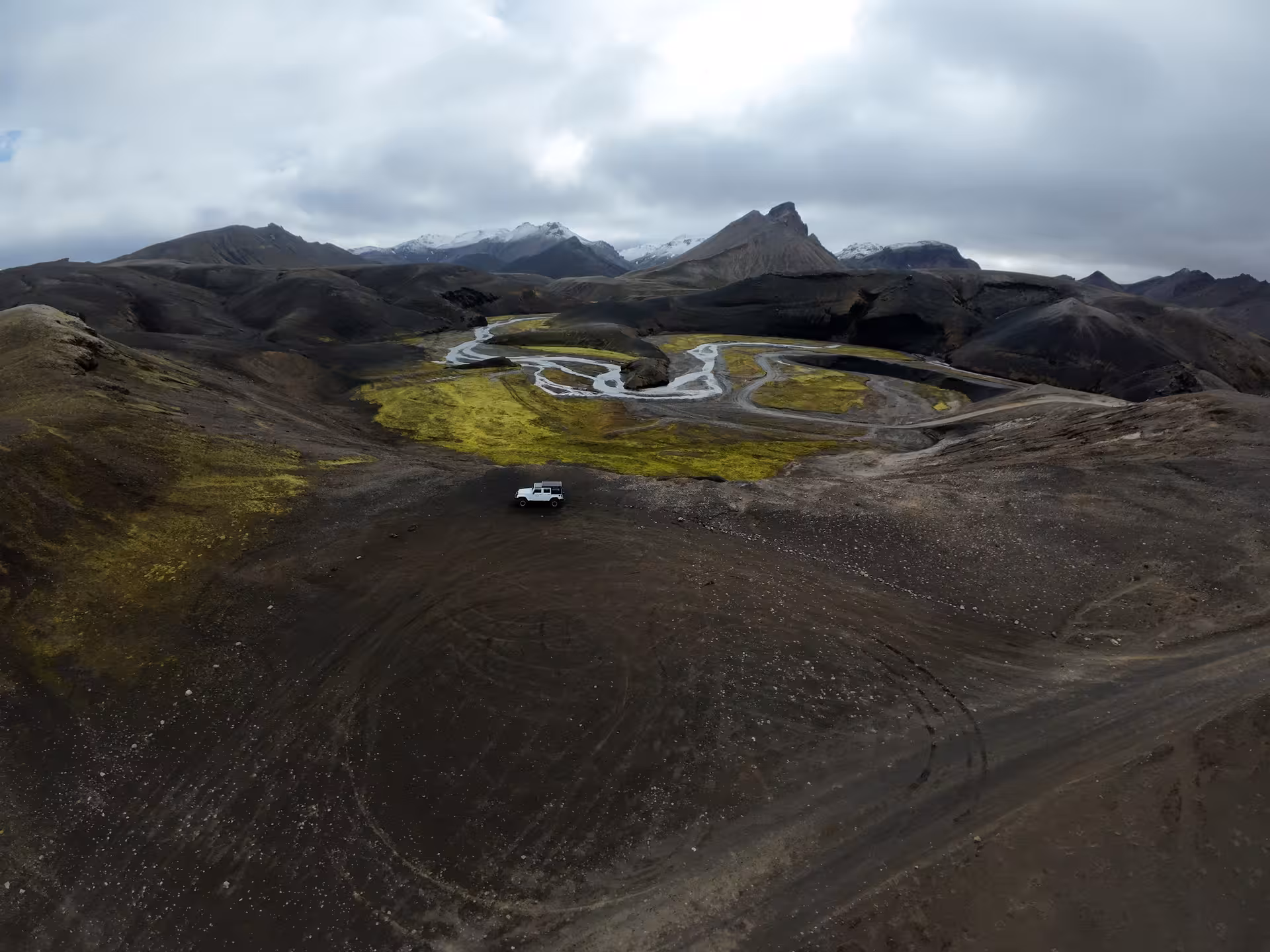 Aerial view of a white Super Jeep on volcanic highland terrain with braided rivers and snow-capped mountains in Landmannalaugar, Iceland