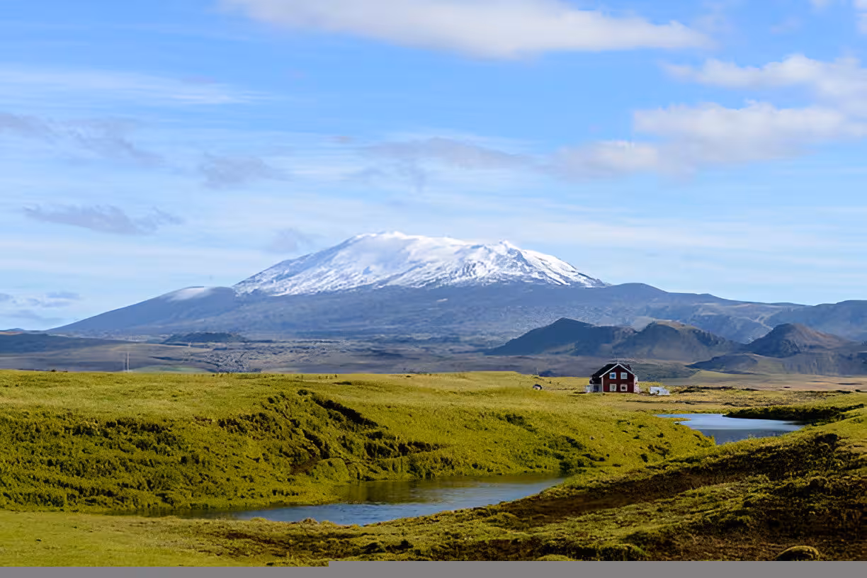 Snow-capped Hekla volcano over green Icelandic highlands with small red farmhouse and pond
