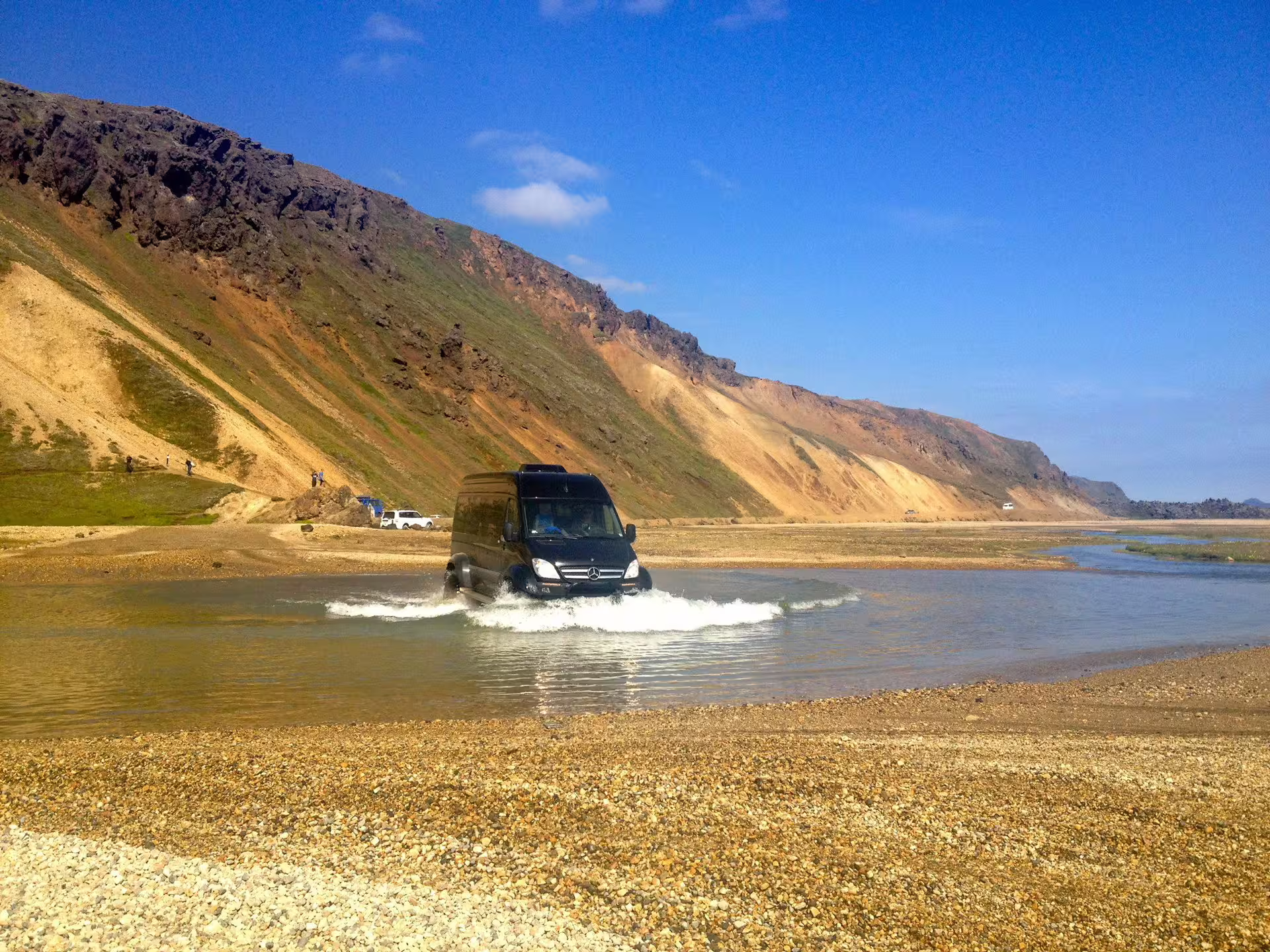 Black Mercedes Sprinter super jeep fording a shallow glacial river in Landmannalaugar Iceland with colorful rhyolite mountains behind