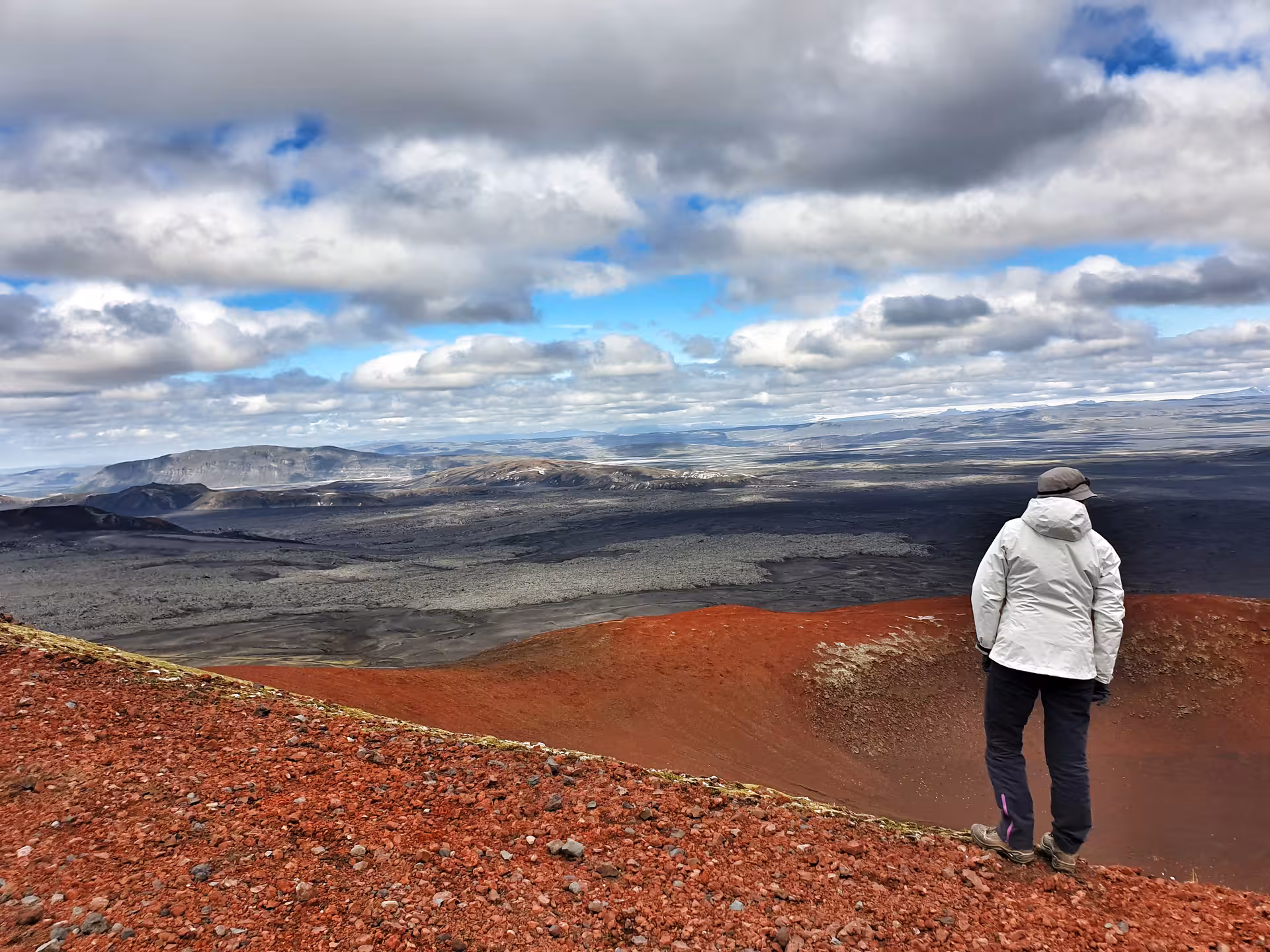 Hiker standing on red volcanic crater rim overlooking vast black lava fields at Landmannalaugar, Iceland