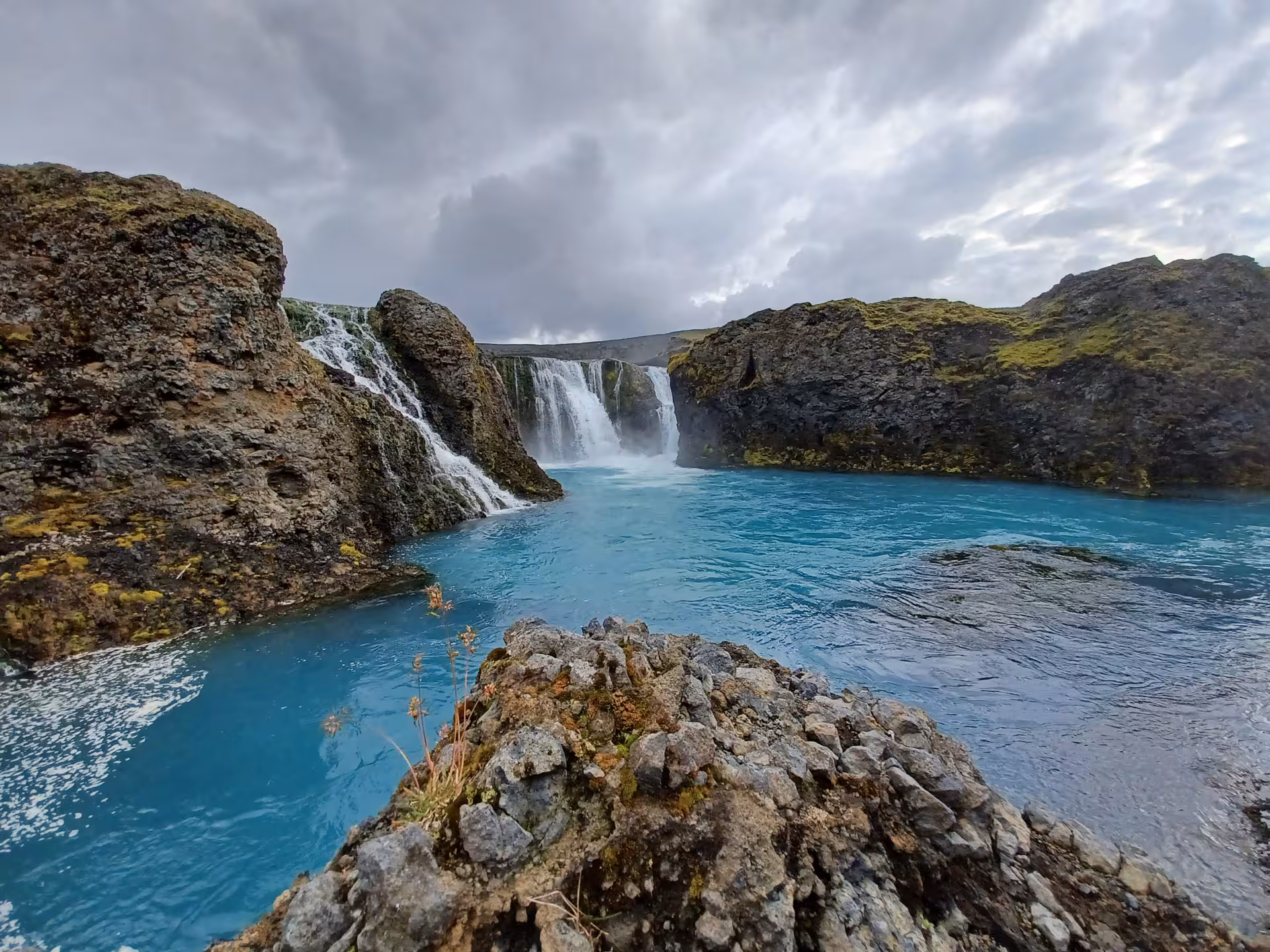 Vivid turquoise river pool below cascading waterfalls surrounded by mossy volcanic rocks in Landmannalaugar, Iceland
