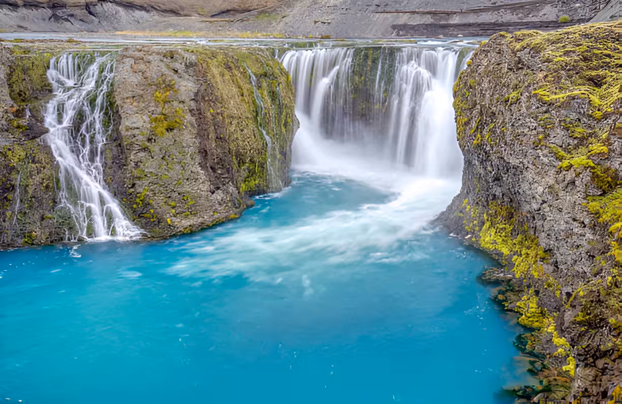 Turquoise glacial river with cascading waterfalls flowing through mossy volcanic rock canyon in Iceland