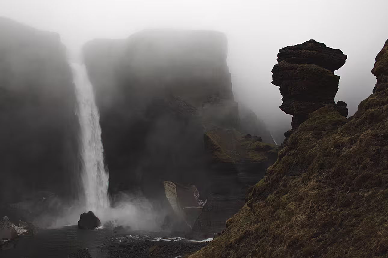 Misty waterfall cascading down dark volcanic cliffs with layered rock formations in Landmannalaugar, Iceland