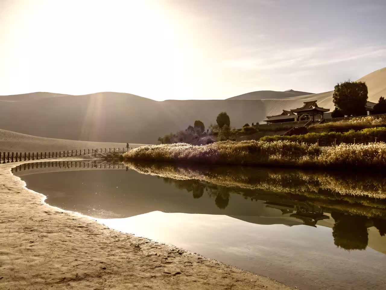 Serene view of Crescent Lake oasis near Lanzhou with traditional Chinese architecture and desert landscape at sunset.
