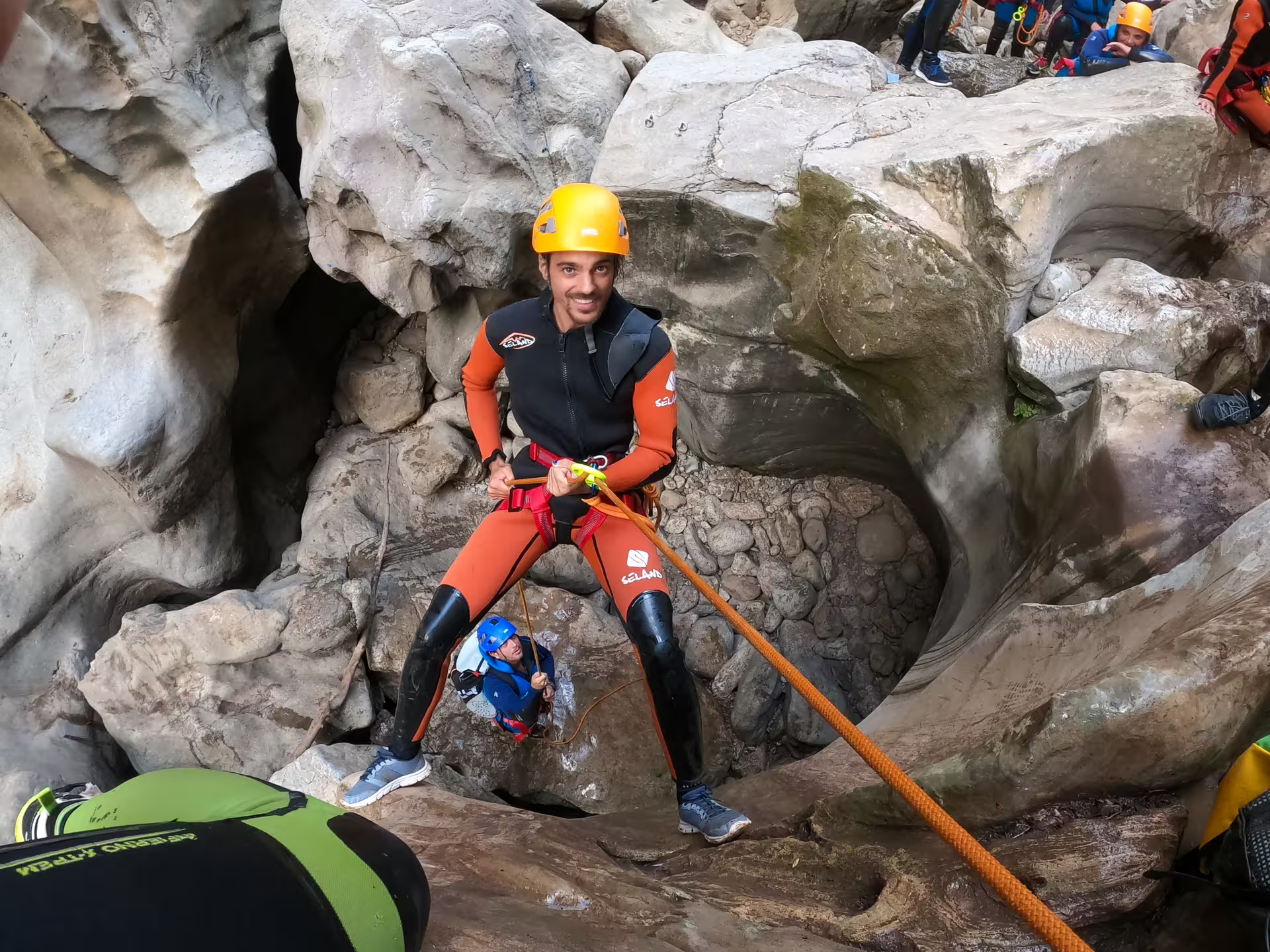 A smiling guide in an orange wetsuit and yellow helmet rappels down rocky canyon walls at Las Buitreras, Málaga.