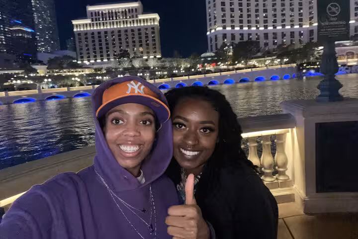 Two smiling women taking a selfie at the Las Vegas Strip at night with the Bellagio fountains behind them