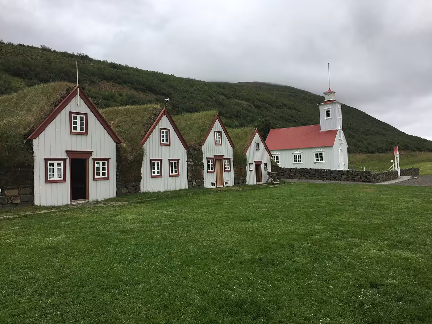Historic white turf houses and red-roofed church at Laufas Museum, Iceland, with green hillside backdrop