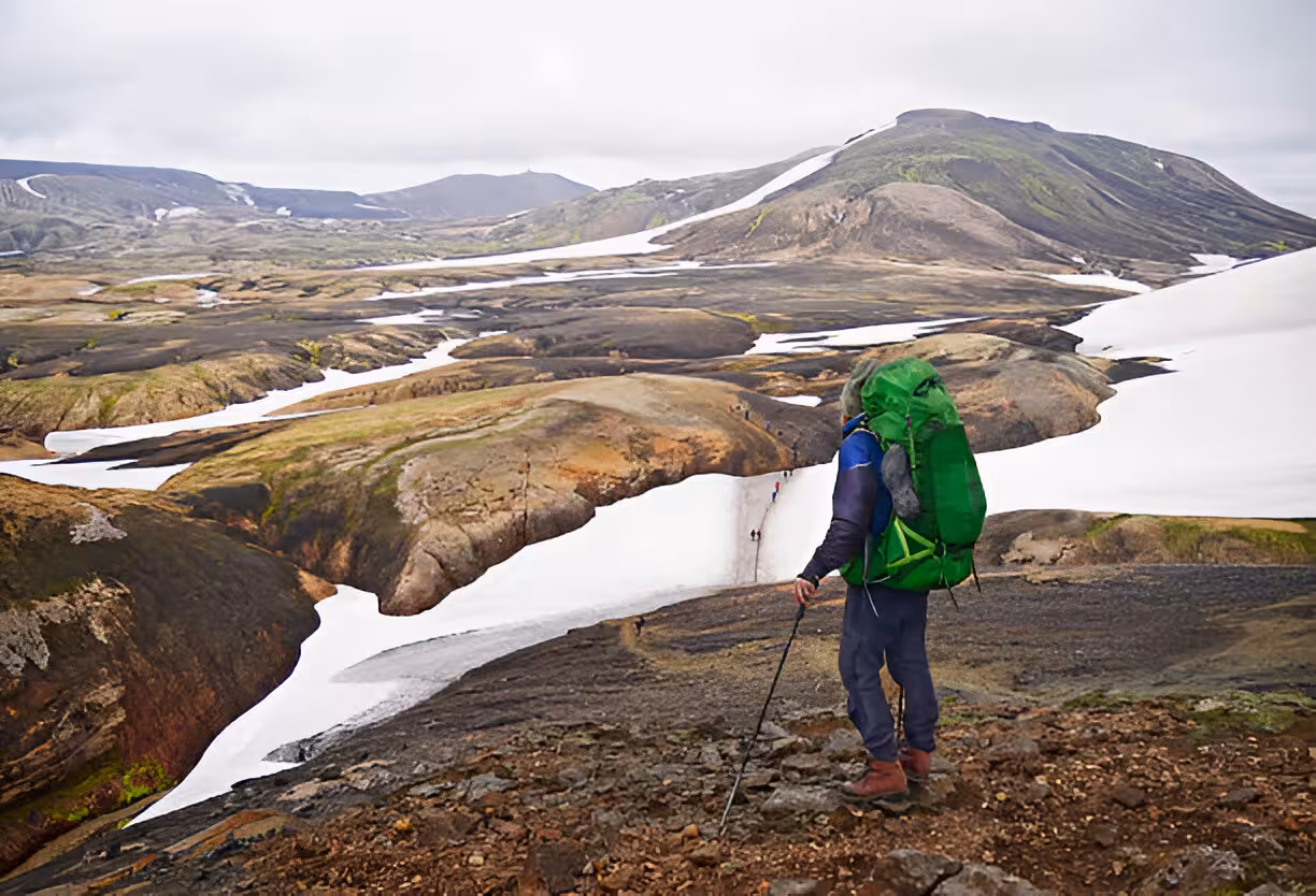 Backpacker with green pack and trekking pole overlooks snowy Laugavegur Trail valley in Iceland