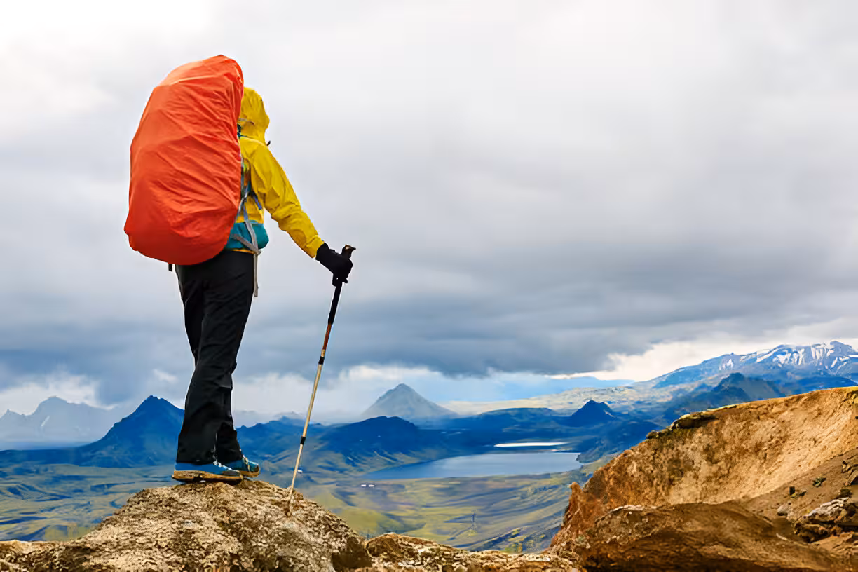 Hiker with red pack cover and trekking pole stands on rocky summit overlooking Iceland's mountains and lake