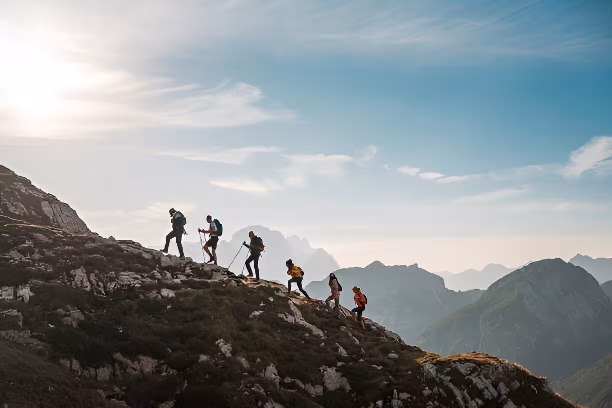 Group of hikers with backpacks and trekking poles ascending a rocky mountain ridge under blue sky