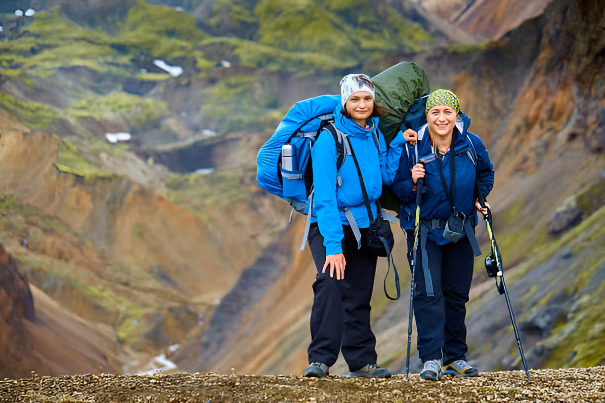 Two female hikers with large backpacks smiling on Laugavegur Trail with colorful Iceland mountains behind