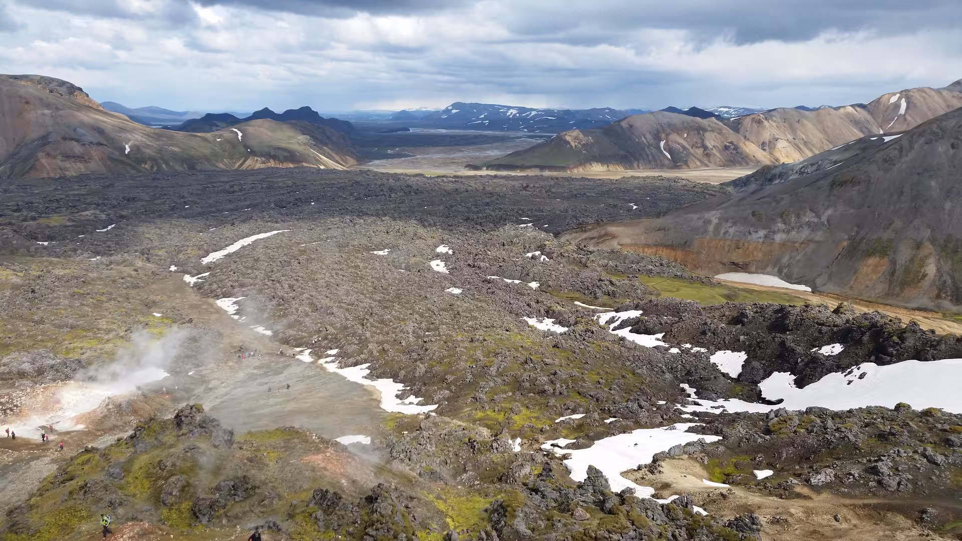 Aerial view of Landmannalaugar lava fields with geothermal steam, snow patches, and colorful rhyolite mountains in Iceland