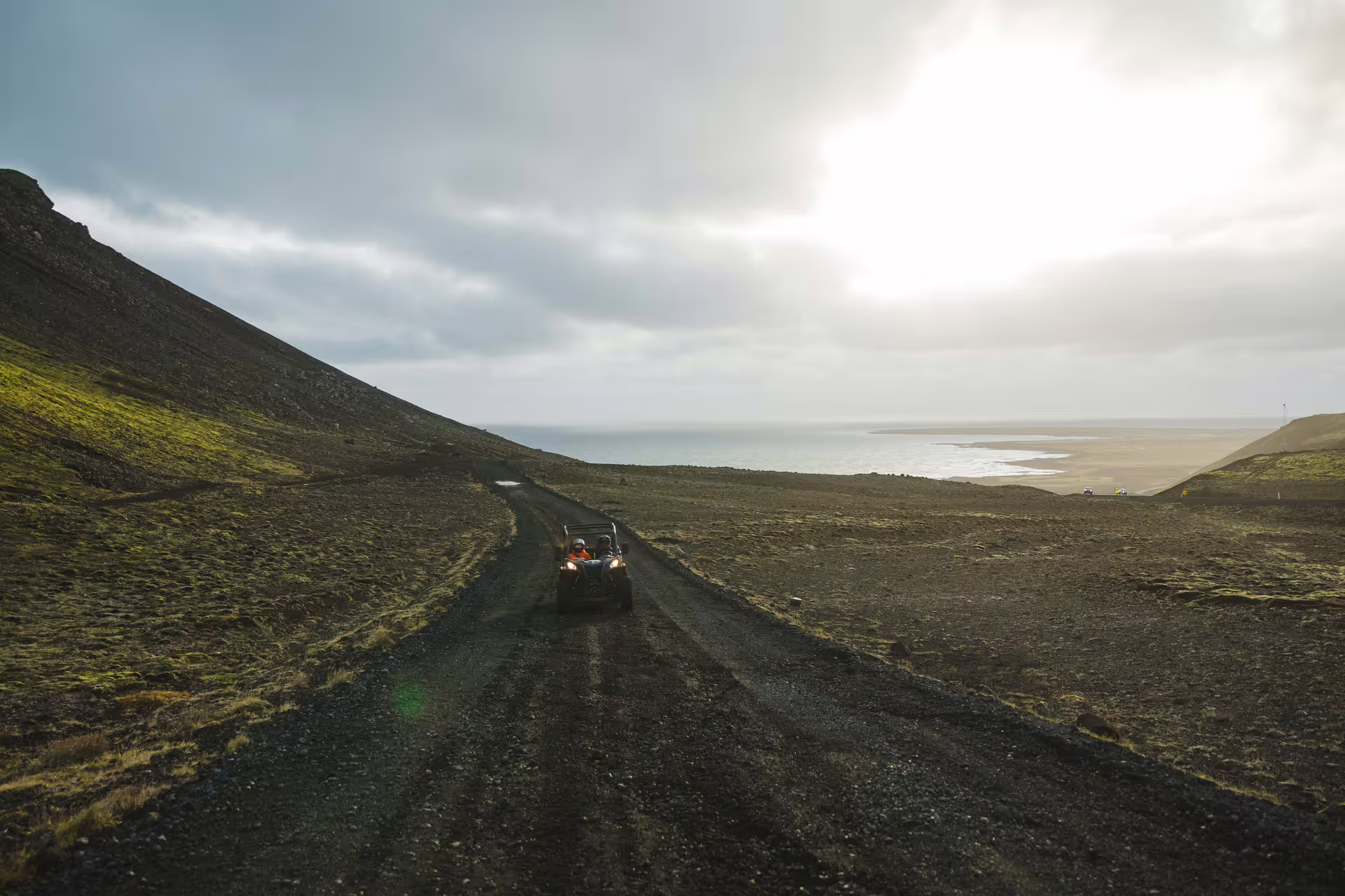 Off-road buggy with headlights on driving down a gravel trail along Iceland's lava coastline under cloudy skies