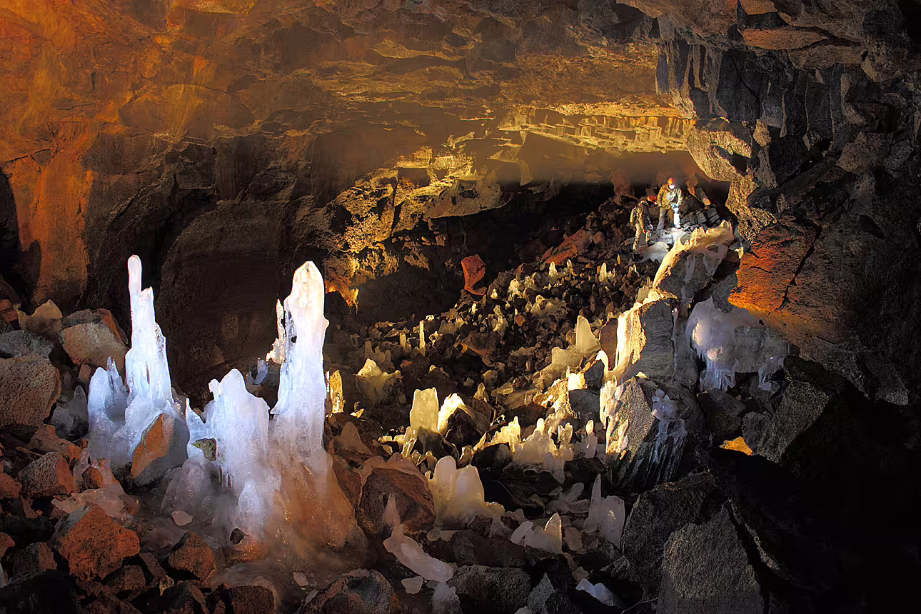 Ice stalagmites and rock formations inside illuminated lava cave tunnel with explorer in background