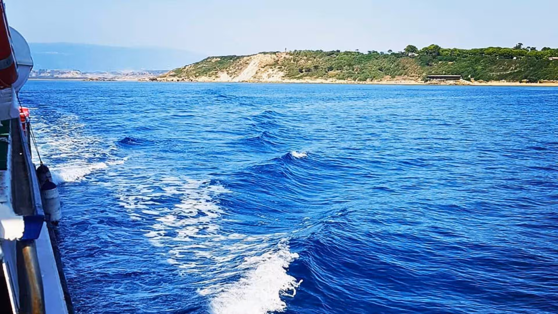 A boat navigates the vibrant blue waters near Capo Rizzuto, with scenic coastal views and gentle waves.