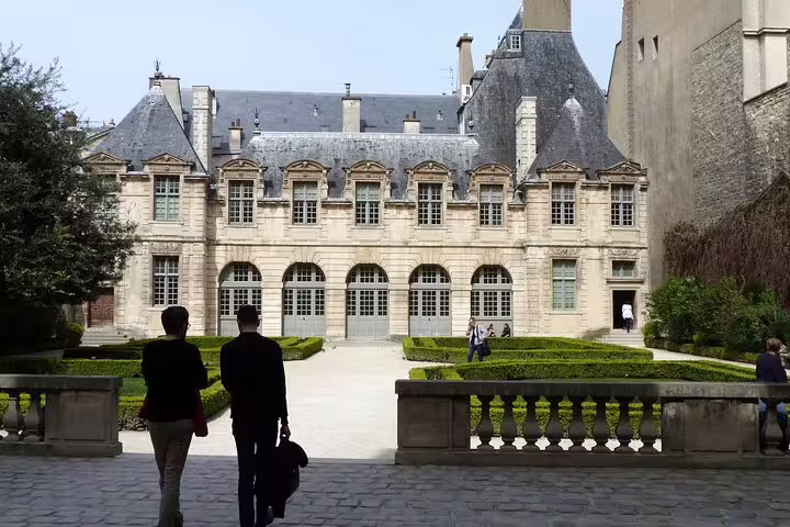 Visitors walking toward a grand historic mansion in Le Marais district, Paris, with manicured hedges.