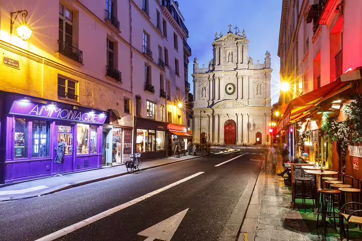 Illuminated Parisian street in Le Marais with glowing shops and historic church facade at dusk