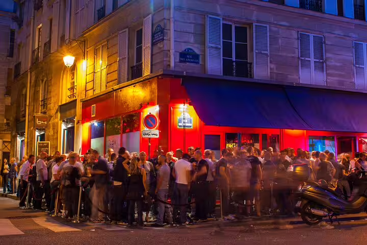 Crowd of people gathered outside a vibrant red-lit bar on a lively Paris street corner at night