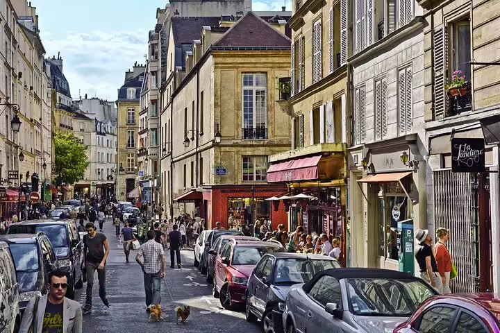 Busy cobblestone street in Le Marais Paris with historic buildings, cafes, parked cars, and pedestrians walking