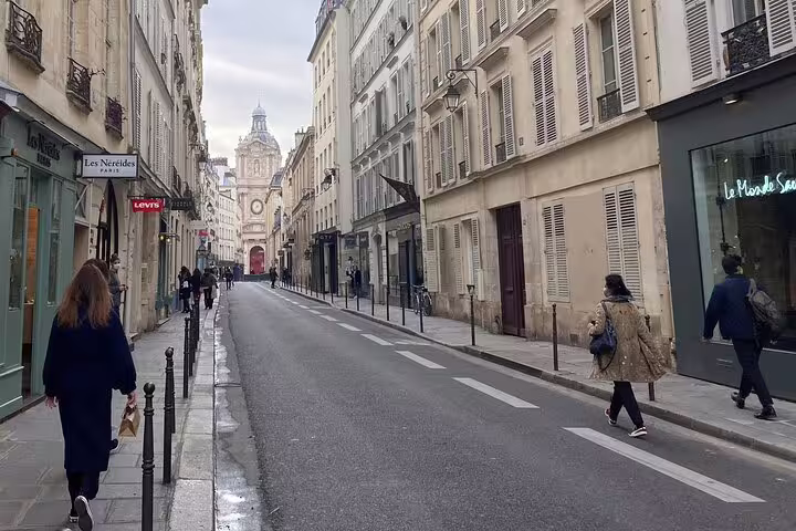 Quiet Parisian street in Le Marais with historic Baroque church at end, pedestrians walking past boutique shops