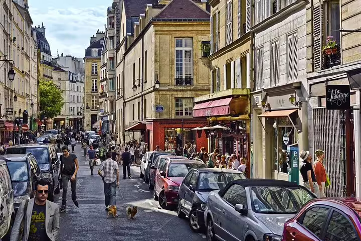 Charming Parisian street in Le Marais with historic buildings, cafes, parked cars, and pedestrians walking.