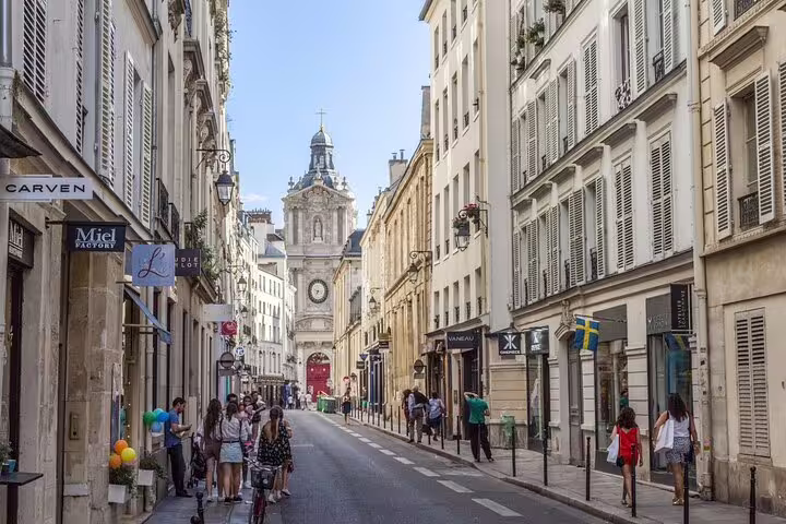Charming Le Marais street in Paris with historic church tower, boutique shops, and pedestrians on a sunny day