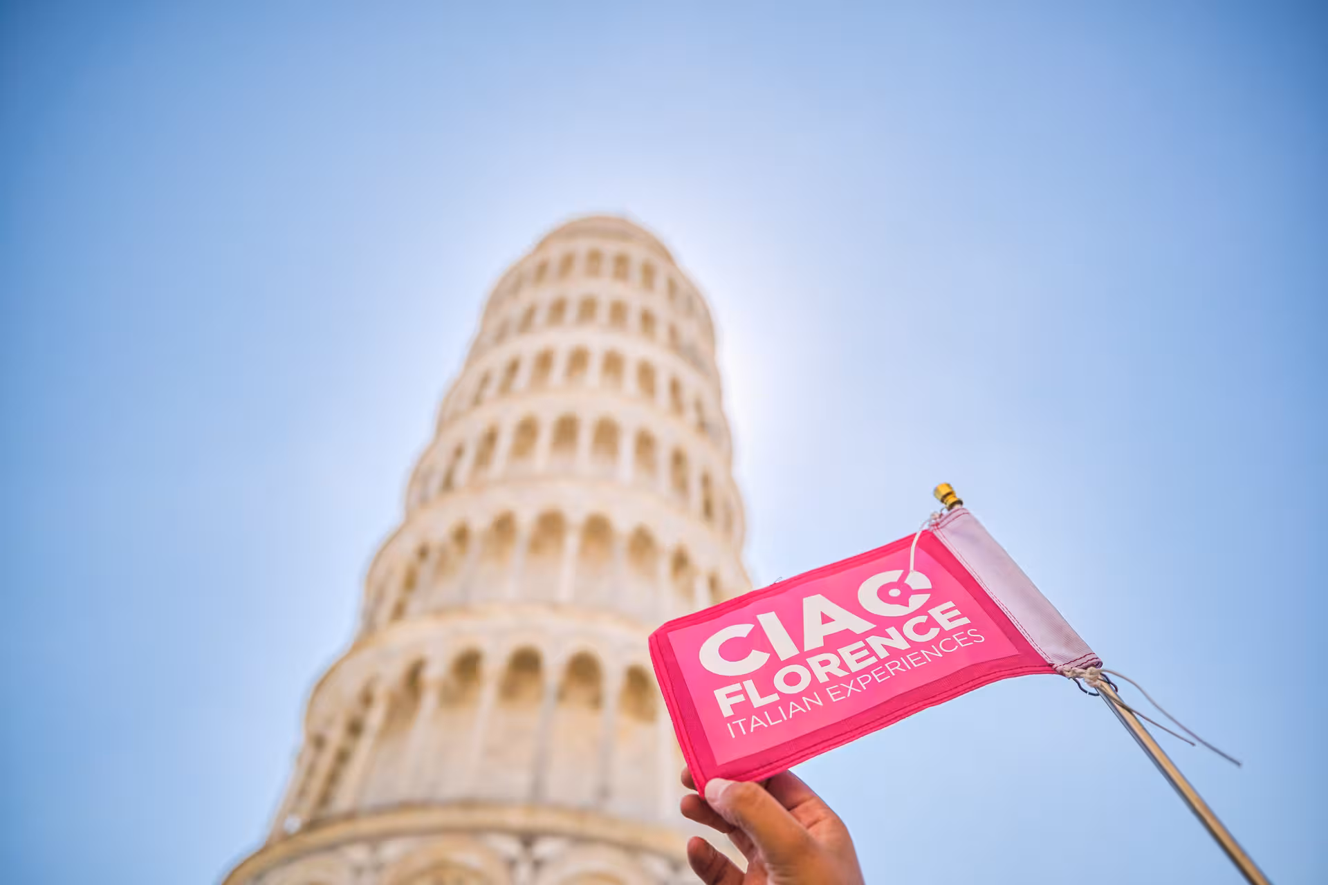 Hand holding a flag in front of the Leaning Tower of Pisa on a clear day during a Florence to Pisa tour.