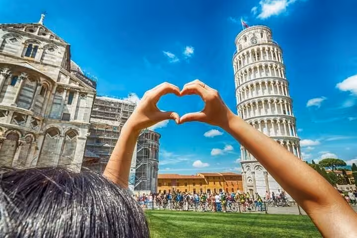 Hands forming a heart shape in front of the iconic Leaning Tower of Pisa under a bright blue sky.