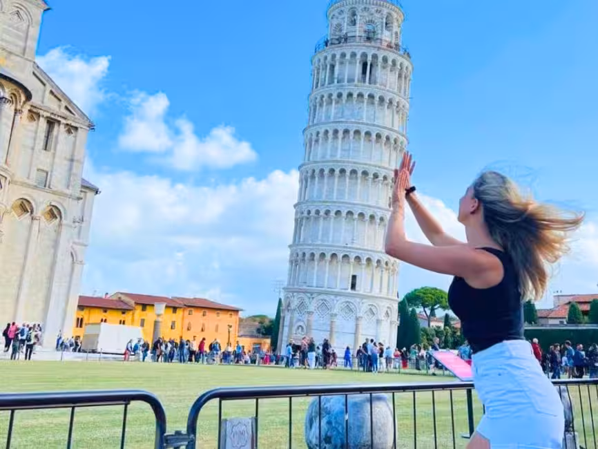 A visitor playfully posing with the iconic Leaning Tower of Pisa under a clear blue sky.