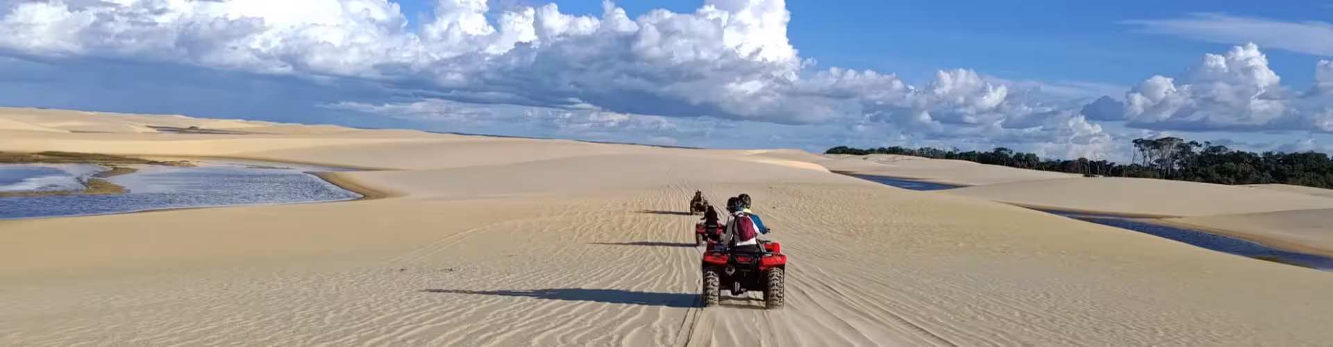Adventure seekers riding red ATVs quad bikes across vast golden sand dunes with scenic lagoons and lush forest under a dramatic blue sky on an exciting off-road quad tour experience.