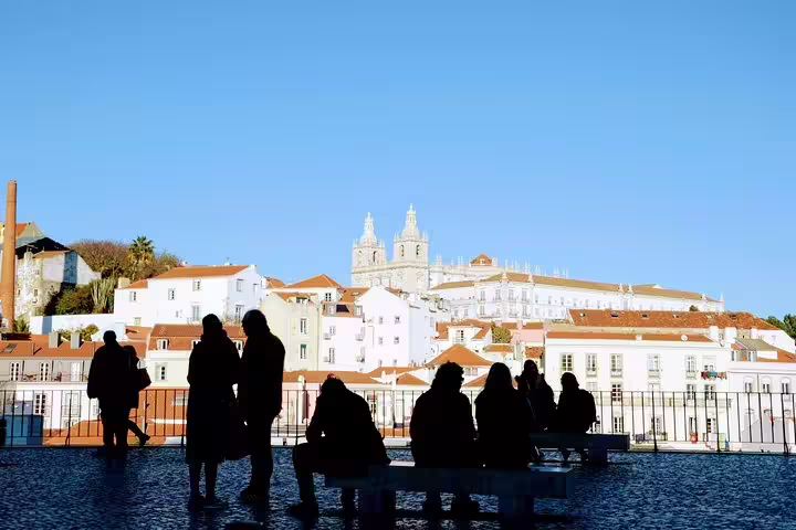 Silhouetted tourists enjoy a panoramic view of Lisbon's historic Alfama district with iconic architecture against a clear blue sky.