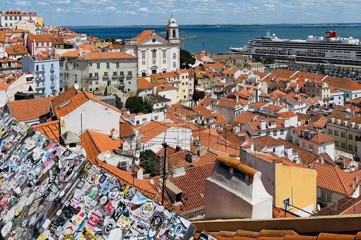 Panoramic view of Lisbon's Alfama district with red rooftops, a cruise ship, and the Tagus River in the background.