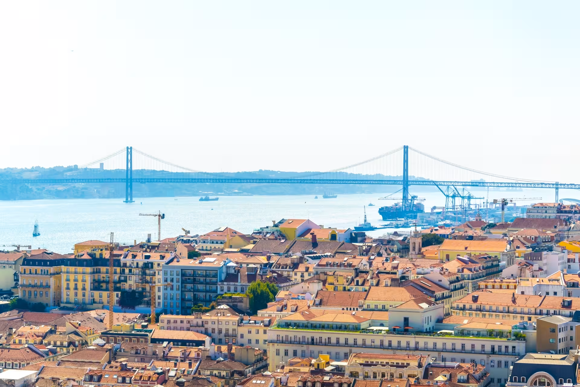Panoramic aerial view of Lisbon old town rooftops with the 25 de Abril Bridge and Tagus River backdrop.
