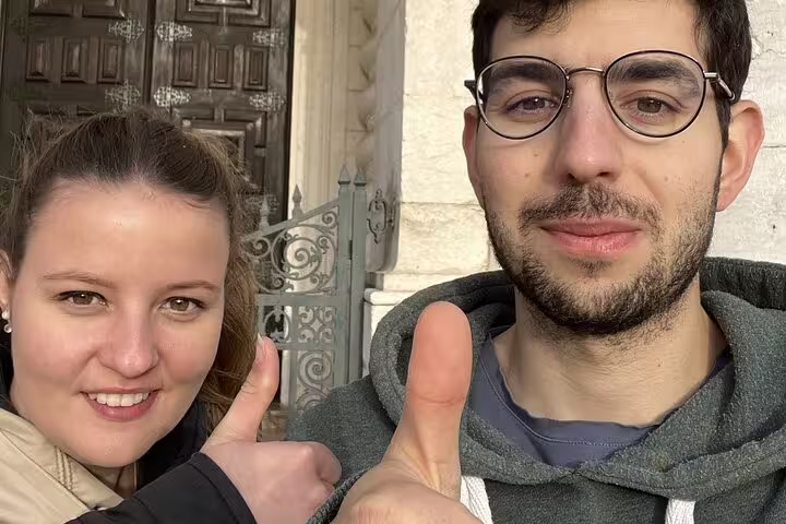 Two smiling tourists giving thumbs up in front of historic Lisbon Alfama building entrance