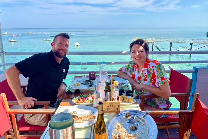 Couple enjoying a scenic seaside lunch with wine on the Setúbal & Arrábida Wine Tour from Lisbon, featuring stunning ocean views.