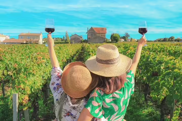 Two women in sun hats toast with wine glasses amidst lush vineyards on the Setúbal & Arrábida Wine Tour from Lisbon.