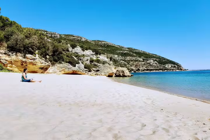 Person relaxing on a pristine beach with clear blue waters and scenic cliffs, part of Lisbon's South Sights & Wine Tour.