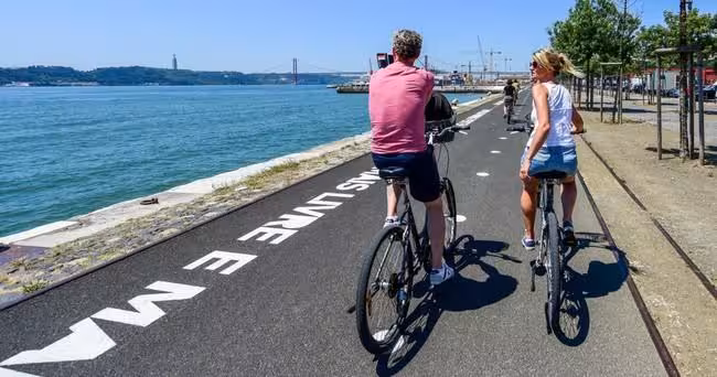 Cyclists riding along Lisbon's scenic waterfront bike path with views of the iconic 25th April Bridge.