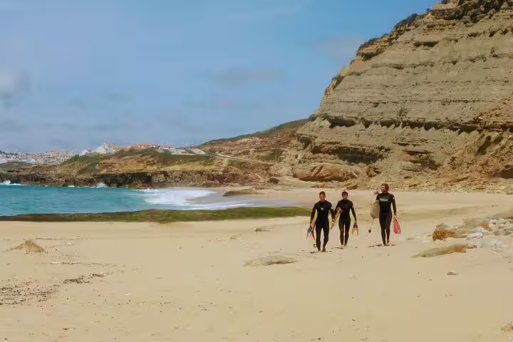 Three people in wetsuits walk along a scenic beach with cliffs, ready for a Lisbon bodyboarding adventure under clear blue skies.
