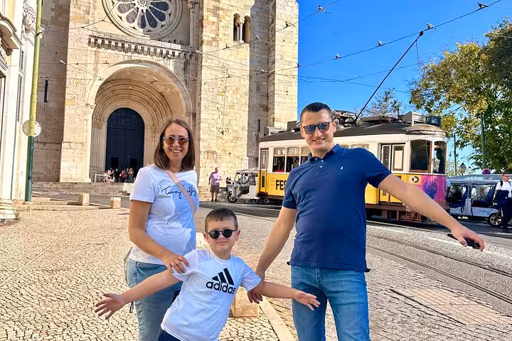 Family posing in front of Lisbon Cathedral with a traditional tram passing by on a sunny day.