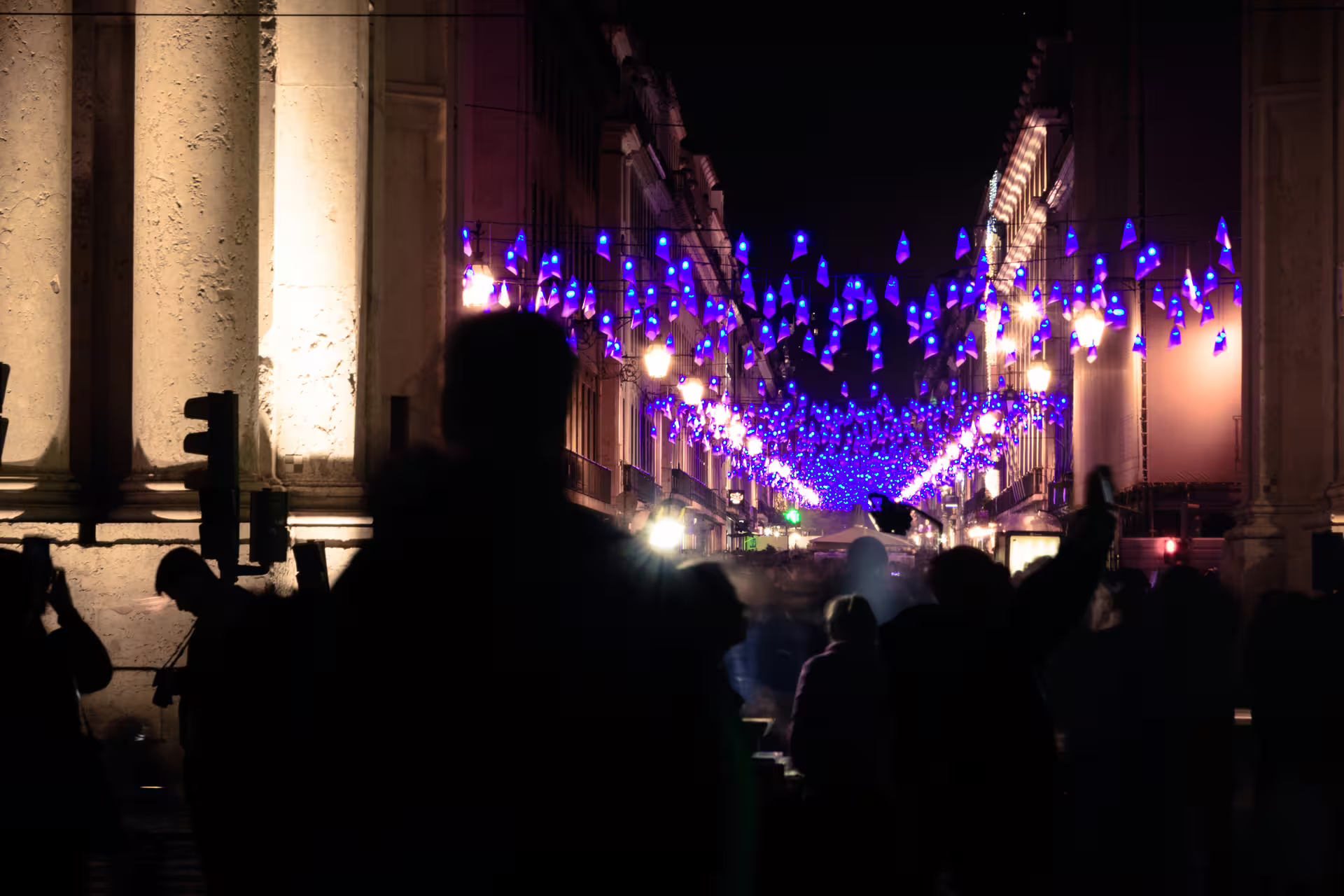 Silhouetted people admire stunning blue Christmas lights illuminating a historic Lisbon street during a festive night walk.