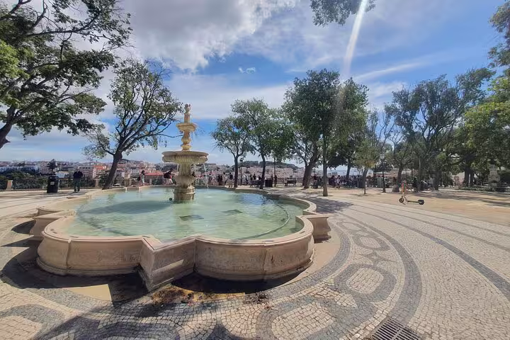 Scenic view of Lisbon's Old Town park with a historic fountain, surrounded by lush trees and intricate mosaic pathways.