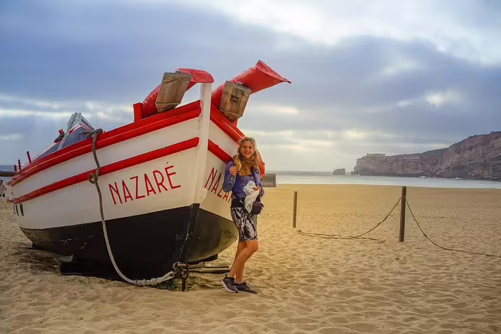 Woman standing beside traditional fishing boat on Nazaré beach, Portugal, with scenic coastal backdrop on Lisbon to Porto tour.