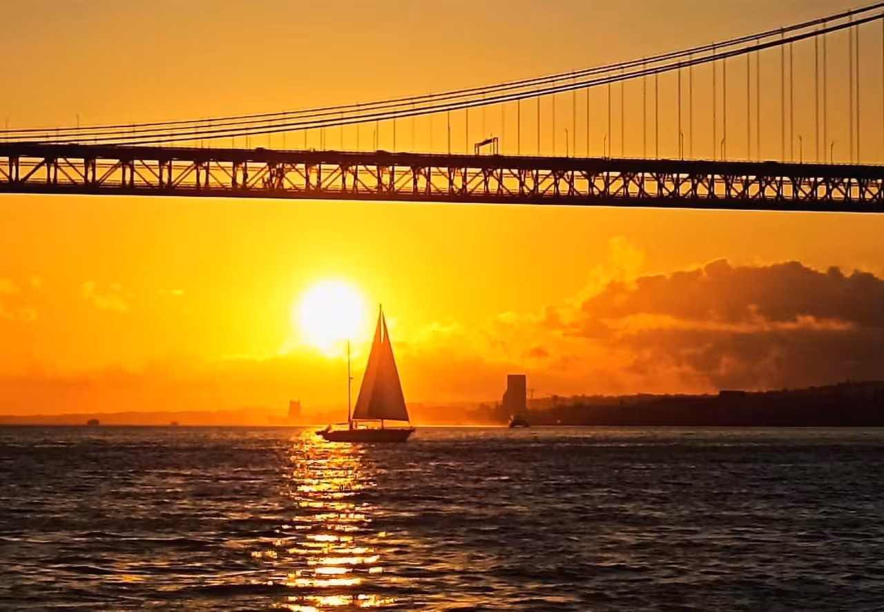 Sailboat gliding under Lisbon’s 25 de Abril Bridge at golden hour on a shared Tagus River sunset cruise