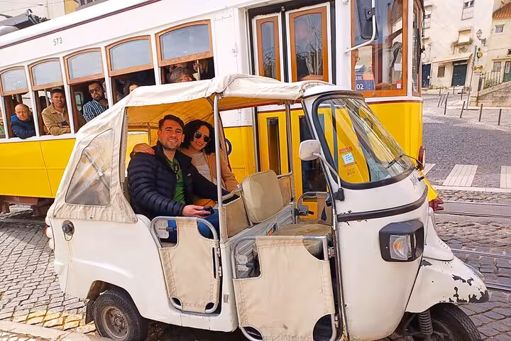 Couple riding a Lisbon tuk tuk beside the yellow Tram 28 in Alfama on a 1-hour Old Town tour